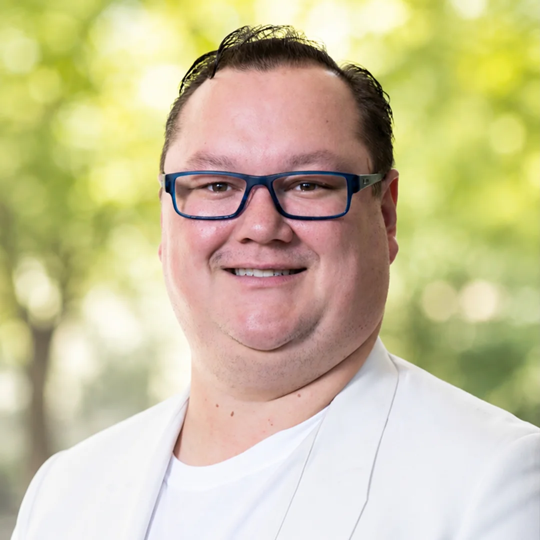 Professional headshot of Matthew smiling outdoors while wearing blue glasses and a light-colored blazer.
